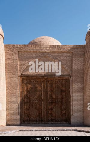 Kunstvoll geschnitztes Holztor in einer alten Backsteinmauer und einem bogenförmigen Portal in Chiwa, Usbekistan. Historische Silk Road Architektur unter klarem blauen Himmel, Trav Stockfoto
