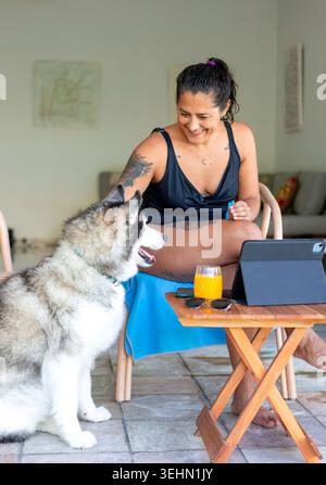 Die lächelnde Frau macht eine Pause von der Fernarbeit zu Hause, verwendet ein Tablet und Smartphone, während sie sich auf einem Stuhl mit einem Husky in der Nähe und einem Glas Saft entspannt Stockfoto