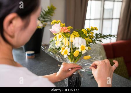 Nahaufnahme einer Frau, die zu Hause einen frischen Frühlingsstrauß einpackt und ein kreatives Hobby und einen ruhigen Lebensstil genießt. Bunte Blumen und Hände, die Zellophan binden Stockfoto