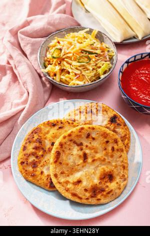 Pupusas auf einem blauen Teller mit Curtido, Salsa roja, Yuca und Humitas auf rosa Hintergrund, die eine traditionelle salvadorianische Mahlzeit und Zentralamerika zeigen Stockfoto