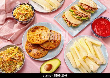 Tischbild mit lateinamerikanischem Familienessen mit Pupusas, Arepas, Curtido, Tajadas, Humitas, Yuca und Salsa auf einem farbenfrohen rosafarbenen Tisch, Tradition Stockfoto