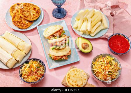 Tafelbild lateinamerikanischer Gerichte von oben auf einem farbenfrohen rosa Tisch: Pupusas, Arepas, Curtido, Tajadas, Humitas, Yuca und Salsa, Set für eine Familie Stockfoto