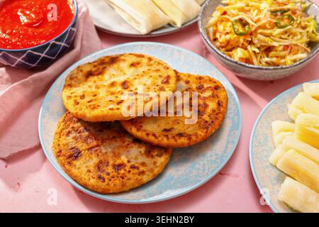 Pupusas auf einem blauen Teller serviert mit Curtido, Salsa roja, Yuca und Humitas auf rosa Hintergrund, die ein traditionelles salvadorianisches Essen und Central A zeigen Stockfoto
