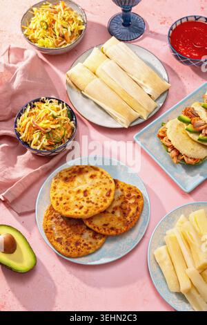 Tafelbild mit lateinamerikanischen Gerichten von oben mit Pupusas, Arepas, Curtido, Tajadas, Humitas, Yuca und Salsa auf einem farbenfrohen rosafarbenen Tisch für Familienessen Stockfoto