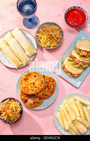 Tafelbild mit lateinamerikanischen Gerichten von oben mit Pupusas, Arepas, Curtido, Tajadas, Humitas, Yuca und Salsa auf einem farbenfrohen rosafarbenen Tisch, Familienessen Stockfoto