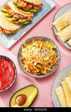 Tafelbild mit Blick von oben auf lateinamerikanische Gerichte mit Pupusas, Arepas, Curtido, Humitas, Yuca und Salsa auf einem farbenfrohen rosafarbenen Tisch, für ein Familienessen. Stockfoto