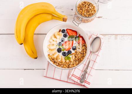 Blick von oben auf eine gesunde Frühstücksschale mit Joghurt, Müsli-Müsli, Bananenscheiben, Erdbeeren und Heidelbeeren, garniert mit Nüssen auf einem weißen Holztisch Stockfoto