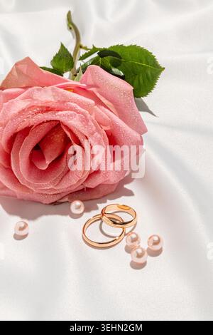 Nahaufnahme des Stilllebens einer rosa Rose mit Tau auf weißem Brautschleier, mit zwei goldenen Trauringen und verstreuten Perlen, die Liebe symbolisieren Stockfoto