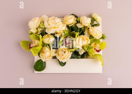 Blick von oben auf ein elegantes Blumenarrangement mit weißen Rosen und grünen Orchideen in einer weißen Geschenkbox auf pastellfarbenem Hintergrund, ideal für Hochzeit, Begrüßung, Stockfoto