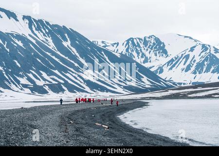 Svalbard und Jan Mayen - 02. Juli 2025: Blick auf eine Gruppe von Touristen in roten Jacken, die an einem dunklen Kiesstrand mit schneebedeckten Bergen und Ruhe stehen Stockfoto