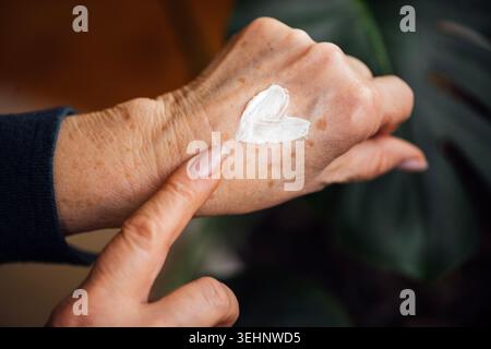 Nahaufnahme einer Hand, die einen herzförmigen Klecks Creme auf das Handgelenk aufträgt, mit einer verschwommenen grünen Pflanze im Hintergrund, die das natürliche Ambiente unterstreicht Stockfoto