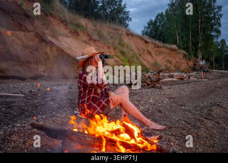 Frau mit Strohhut und kariertem Hemd sitzt barfuß am Lagerfeuer an einem Sandstrand in der Abenddämmerung und durchsucht die natürliche Landschaft mit einem Fernglas während eines friedlichen Outdoor-Campingausflugs Stockfoto