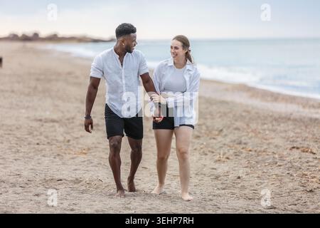Ein Paar genießt einen gemütlichen Spaziergang am ruhigen Strand von Zapillo in Almeria, Spanien. Sie laufen barfuß auf dem warmen Sand, halten Hände und teilen sich Stockfoto