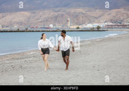 Ein Paar genießt einen gemütlichen Spaziergang entlang der Sandküste von Playa del Zapillo in Almeria, Spanien, wobei er Hände hält und die Sonne genießt. Das klare Wasser Stockfoto