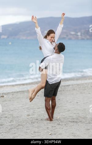 Ein Paar genießt einen freudigen Moment an den Sandstränden von Playa del Zapillo in Almeria, Spanien. Die Wellen des Meeres schlagen sanft in der Nähe, und der Mann hebt sich Stockfoto