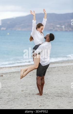 Ein Paar genießt einen Moment der Freude am Zapillo Strand in Almeria, Spanien. Der Mann hebt die Frau in die Luft, während sie aufgeregt ihre Arme hebt, was einen erzeugt Stockfoto