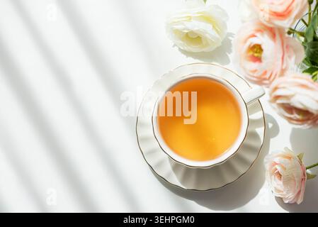Elegante Lifestyle-Komposition mit weißer Keramik-Teetasse mit Muscheluntertasse und goldenem Rand, gefüllt mit bernsteinfarbenem grünem Tee, umgeben von blassrosa Stockfoto