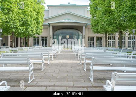 Symmetrischer Blick auf leere weiße Holzbänke, die für ein Konzert im Freien vor dem Musikpavillon angeordnet sind. Grüne Bäume und Bürgersteig in einem ruhigen Spa Stockfoto