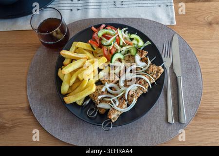 Blick von oben auf gegrillte Schweinefleisch-Souvlaki-Spieße, serviert mit Pommes frites, Salat und Zwiebeln auf einem schwarzen Teller. Stockfoto