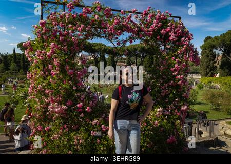 Rom, Italien - 24. Mai 2026: Eine junge Frau mit Sonnenbrille und schwarzem Kaktus-T-Shirt posiert lächelnd vor einem großen Herzbogen, der mit bl bedeckt ist Stockfoto
