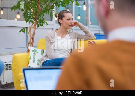 Eine mittlere Aufnahme, die sich auf eine Frau in einer beigen Strickjacke konzentriert, die auf einem gelben Bürosofa sitzt. Sie hört einem Kollegen aufmerksam zu (verschwimmt im Voraus Stockfoto