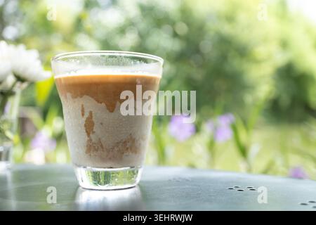 Dreckiger Kaffee mit Espresso, der über Milchschaum in klarem Glas auf dem Tisch im Freien mit violetten Wildblumen-Bokeh-Hintergrund-Gartencafé gegossen wurde Stockfoto