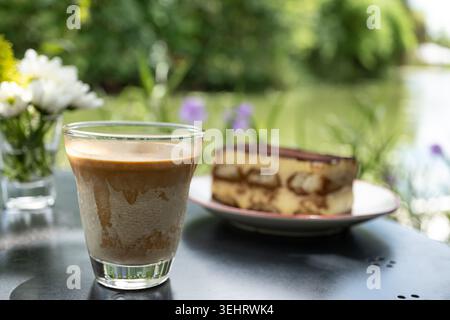 Schmutziges Kaffeeglas mit Tiramisu-Kuchenscheibe im Hintergrund im tropischen Gartencafé im Freien mit Blumen und Blick auf den Fluss in Thailand Stockfoto