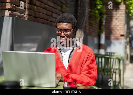 Konzentrierter positiver afroamerikaner, der draußen im Straßencafé in der Stadt am Laptop arbeitet. Stockfoto