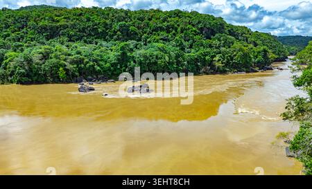 Der Guayabero River fließt durch einen üppig grünen Wald mit großen Felsen, die aus schlammigen Gewässern unter teilweise bewölktem Himmel hervorgehen. Stockfoto