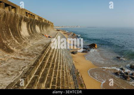 Weite Küstenlandschaft mit einer großen gebogenen Betonmauer, Sandstrand, felsiger Küste und ruhigem Ozeanwasser unter der warmen Abendsonne. Eine Person Stockfoto
