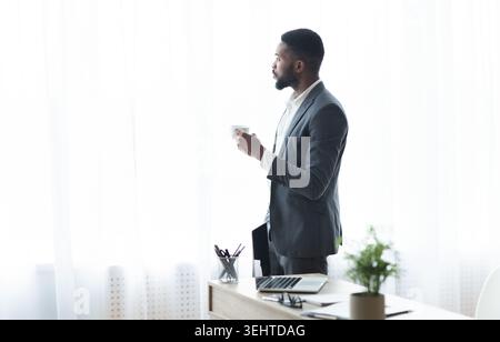 Nachdenklicher schwarzer Geschäftsmann, der aus dem Fenster blickt und Kaffee trinkt Stockfoto