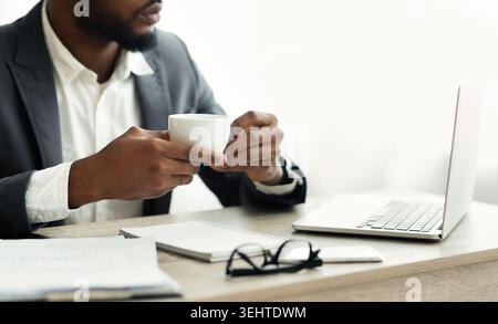 Mann in Anzug genießt Kaffee, während er an einem Schreibtisch an einem ruhigen Morgen am Laptop arbeitet Stockfoto