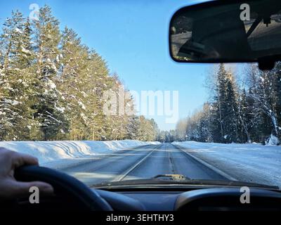 Eine Person fährt auf einer verschneiten Straße mit Sonnenlicht durch die Windschutzscheibe. Die Szene zeigt schneebedeckte Bäume entlang der Route. Stockfoto