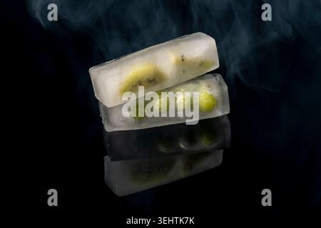 Zwei große Eiswürfel mit Trauben und Kiwischeiben innen auf einer schwarzen reflektierenden Oberfläche und Rauch oben Stockfoto