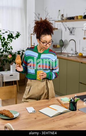 Frau mit lockigem Haar isst ein Croissant und frischen Saft, während sie ihren Tag zu Hause plant. Stockfoto