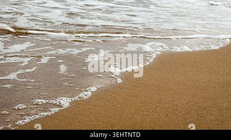 Sanfte Wellen, die gegen einen Sandstrand plätschern, mit Schaum am Wasserrand und einer ruhigen Meeresoberfläche im Hintergrund Stockfoto