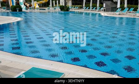 Swimmingpool mit klarem blauem Wasser und gemusterten Fliesen, umgeben von Liegestühlen und Wasserrutschen in einem modernen Erholungsbereich Stockfoto