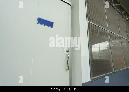 Weiße geschlossene Tür mit Metallgriff und blauem, leerem Typenschild neben einem großen Fenster mit Gittergitter an der Außenfassade des Gebäudes für eine ruhige und ruhige Gebühr Stockfoto