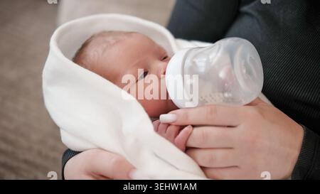 Die Frau Füttert Das Neugeborene Mit Einer Flasche, Während Sie Das Baby Während Des Ruhigen Nachmittags Zu Hause In Ein Tuch Gewickelt Hält. Stockfoto