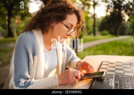 Junge Frau, die mit Smartphone und Laptop aus der Ferne im Park bei Sonnenuntergang arbeitet Stockfoto
