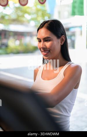 Vertikale mittlere Aufnahme einer glücklichen jungen Kundin Isan Thai, die lächelt und aktiv mit ihrem Arm ausstreckt, um eine Touchscreen-automatische se zu bedienen Stockfoto