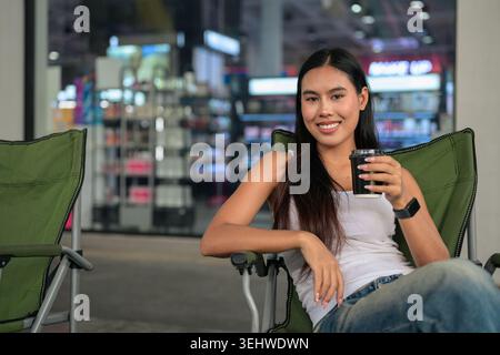 Horizontales mittleres Porträt einer fröhlichen jungen Isan Thai-Frau, die lächelt und direkten Augenkontakt mit der Kamera herstellt, während sie entspannt in einem Grün sitzt Stockfoto