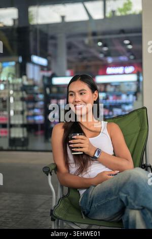 Vertikale mittlere Aufnahme einer glücklichen jungen Isan Thai-Frau, die lächelt und aus der Kamera nach rechts blickt, während sie entspannt in einem grünen, faltbaren Campingkaffee sitzt Stockfoto
