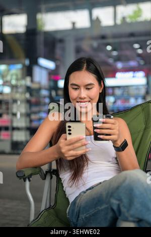 Vertikale mittlere Aufnahme einer glücklichen jungen Kundin von Isan Thai, die nach unten blickt und lächelt, während sie mit ihrem Smartphone interagiert. Entspannt sitzen in einem gr Stockfoto