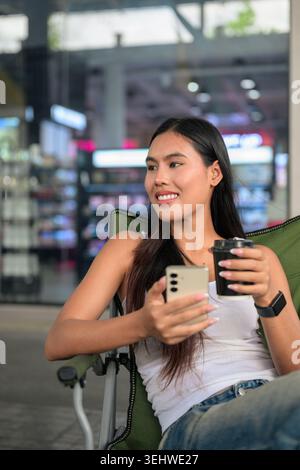 Vertikale Mittelaufnahme einer glücklichen jungen Isan Thai-Konsumentin, die lächelt und aus der Kamera nach rechts blickt. Entspannt in einem grünen Klappcamping sitzen Stockfoto
