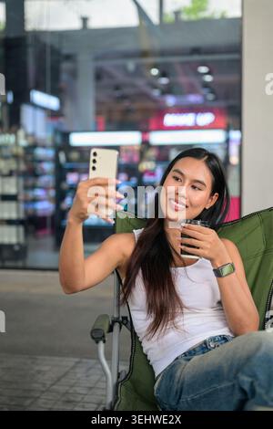 Vertikale mittlere Aufnahme einer glücklichen jungen Isan Thai-Künstlerin oder Influencerin, die ihr Smartphone hochhält, um ein Selfie-Porträt zu machen Stockfoto