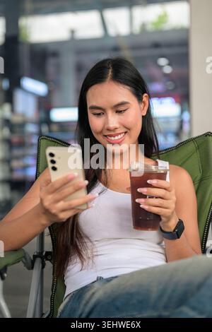 Vertikale mittlere Nahaufnahme einer glücklichen jungen Kundin Isan Thai, die nach unten blickt und lächelt, während sie ihr Smartphone bedient. Entspannt sitzen in A Stockfoto