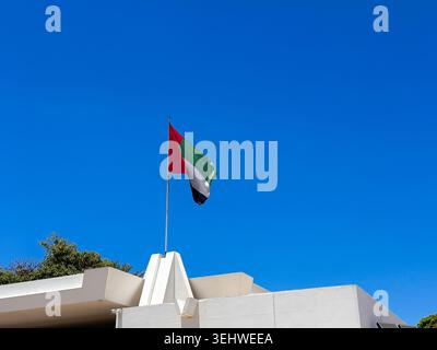 Eine lebendige Flagge der Vereinigten Arabischen Emirate flattert im hellen Sonnenlicht vor einem wolkenlosen blauen Himmel, der auf einer weißen architektonischen Struktur mit weit entfernten Bergrücken montiert ist. Stockfoto
