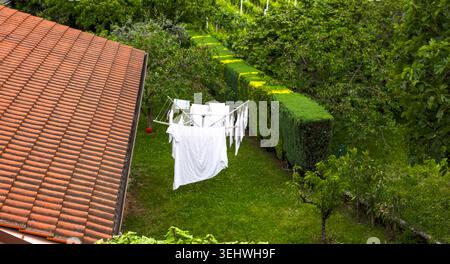 Weiße Kleidung, die auf einer rotierenden Wäscheleine in einem grünen Garten neben einem Haus mit gekacheltem Dach trocknet. Draufsicht. Stockfoto