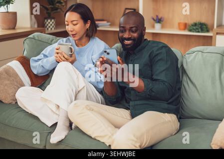 Multiethnische Paare, die sich zu Hause entspannen. Die Frau trinkt Kaffee und der Mann benutzt ein Smartphone. Stockfoto